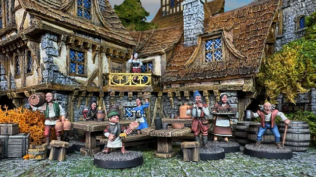 A detailed scene depicts a bustling tavern setting with various miniatures representing villagers and tavern patrons. In the foreground, a young server in a green hat holds a tray, while other characters, including a woman with a basket and a man with mugs, engage in lively activity around wooden tables. The backdrop features a rustic tavern with a thatched roof and stone walls, enhancing the fantasy village atmosphere.