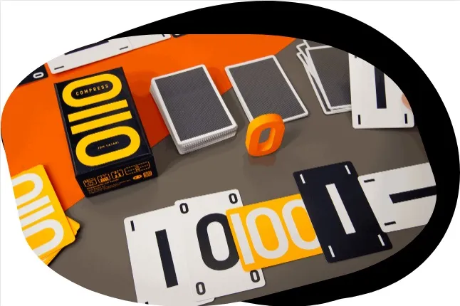 A tabletop setup features the game "Compress," with its distinctive box prominently displayed. Surrounding the box are various playing cards, including black and white cards with binary numbers (1s and 0s) printed on them. An orange game piece shaped like the number "0" is also visible, alongside a couple of decks of standard playing cards. The background includes a vibrant orange surface, enhancing the game's colorful presentation.