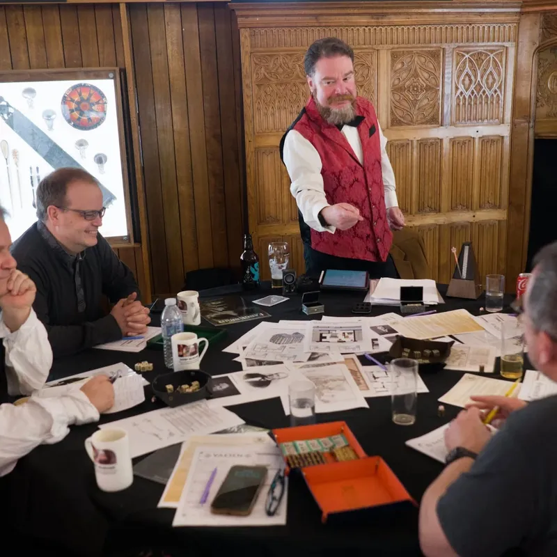 A group of four people is gathered around a table covered with various gaming materials, including character sheets, dice, and notes. One man, dressed in a red vest and black bow tie, stands animatedly, gesturing as he engages the group. The setting features wooden paneling and a warm atmosphere, enhancing the gothic theme of the event.
