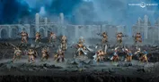 Miniature figures of eight Stormcast Eternals in battle poses, armored in gold and silver, with shields, weapons, and helmets. One figure holds a large hammer, another a staff with a glowing orb. Ruined battlefield with debris and a distant fortress under dark, smoky sky.