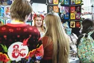 A festive scene at a board game festival features a woman with antlers and floral decorations in her hair, engaging with two attendees. One attendee wears a red sweater adorned with a Dungeons & Dragons logo, while the other has long, straight hair. Behind them, a display showcases various colorful artwork and merchandise, including keyrings for sale.