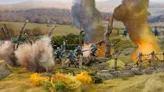 A detailed diorama depicts a World War I battlefield scene with soldiers in blue uniforms engaged in combat amidst smoke and explosions. In the background, a landscape of rolling hills is visible, with barbed wire and trees partially obscured by billowing smoke. The foreground features vibrant yellow foliage, enhancing the dramatic atmosphere of the scene.