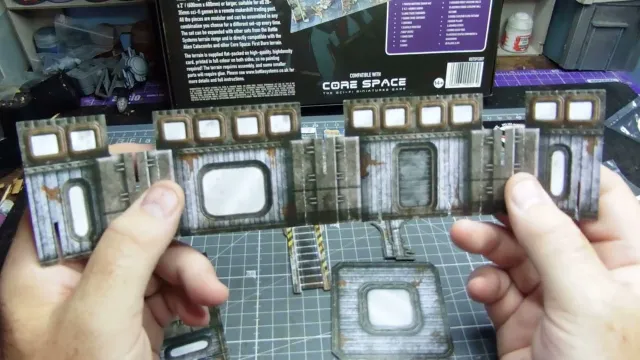 A hand holds a piece of unpainted cardboard terrain with a rectangular window and weathered metal texture. On a green cutting mat, additional assembled cardboard terrain pieces with similar windows, ladders, and industrial details are visible. In the background, a product box for "CORE SPACE" by Battle Systems is partially visible, with text and a barcode.