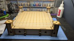 A wooden Rumble Slam Rookie Ring is displayed, featuring a square mat with a grid pattern and four corner posts adorned with star designs. The ring is surrounded by white ropes, and the base has a simple, clean design with additional star motifs. In the background, various paint pots are visible, indicating a workspace for miniature painting and modeling.