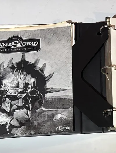 A three-ring binder containing the playtest manuscript for "ManaStorm: The Magic Adventure Game," featuring a black and white cover illustration of a character with a spiked helmet and a weapon. The title "MANASTORM The Magic Adventure Game" is prominently displayed at the top, along with the version number and date on the first page inside the binder. Additional pages include loose character sheets and player handouts, indicating the game's mechanics and spells.