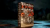 A box for the "Pirate Borg Starter Set" stands prominently, featuring bold, stylized text in red and white against a dark background. The artwork depicts a group of diverse pirate characters, including a skeletal figure, a woman in a dress, and a muscular man, all set in a chaotic, adventurous scene. The box is placed on a wooden surface, with hints of a ship's silhouette and water in the background, enhancing the nautical theme.