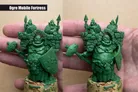 A detailed green sculpt of an "Ogre Mobile Fortress" is held in a person's hand, showcasing a large, rotund ogre character adorned with armor and a shield. The ogre is flanked by two smaller figures, likely goblins, peeking over a stone wall structure on the ogre's back. The sculpt features intricate details, including facial expressions and textures, emphasizing the whimsical and mischievous nature of the characters.