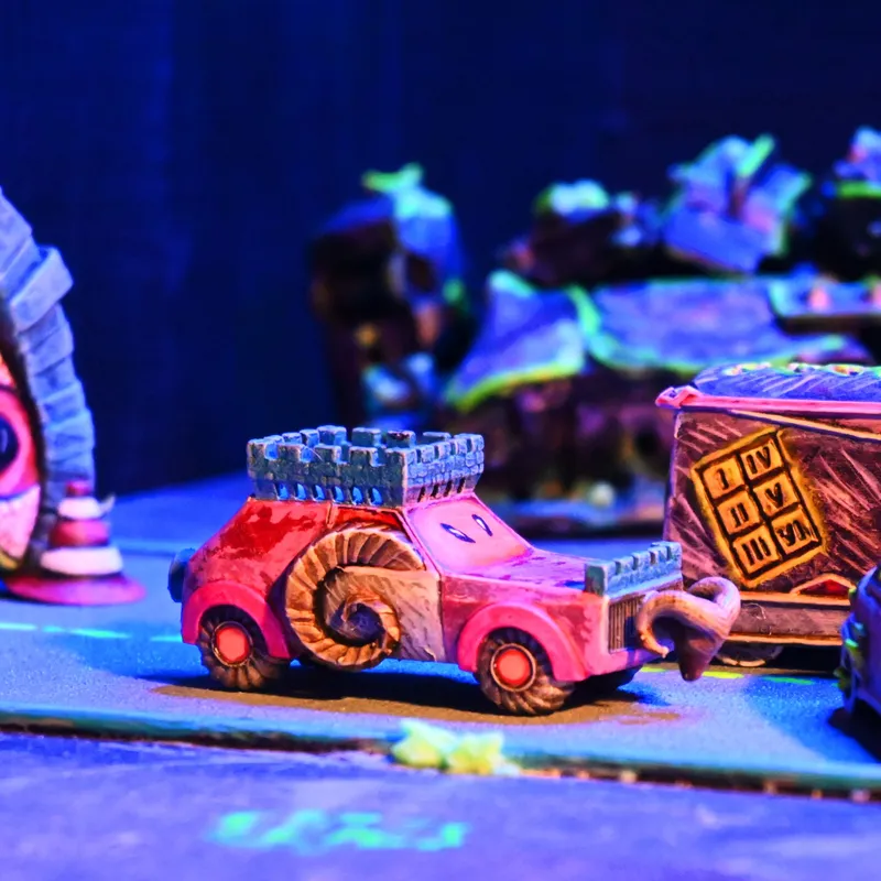 A colorful miniature vehicle designed for a racing game, featuring a pink body with a whimsical spiral design and a castle-like turret on top. In the background, other fantastical vehicles and structures are visible, set against a vibrant, illuminated tabletop environment. The scene captures a playful and chaotic atmosphere, fitting for a greenskin-themed racing skirmish.