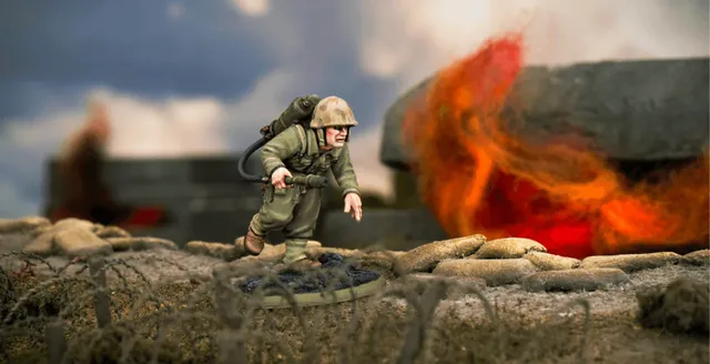 A detailed miniature of a U.S. soldier, Hershel W. 'Woody' Williams, is depicted in action, equipped with a flamethrower on his back and wearing a helmet and military uniform. He is positioned on a textured base that resembles a battlefield, with a backdrop of flames and smoke suggesting a combat environment. The scene captures the intensity of the Pacific theatre during World War II.