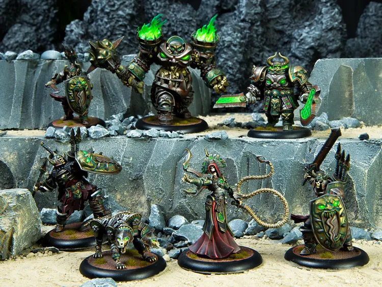 A group of intricately detailed miniatures from the Warmachine tabletop game is displayed on a rocky terrain backdrop. The figures include a large, armored character with green flames emanating from its shoulders, flanked by a menacing wolf-like creature and several other warriors adorned with ornate armor and shields. In the foreground, a female character in a flowing red robe wields a whip-like weapon, while another figure holds a shield and a weapon, showcasing the rich variety of designs and colors typical of the game's factions.