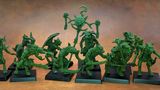 A collection of old school miniatures from Satyr Art Studio, featuring various fantastical creatures in a vibrant green color. The figures include beastmen with distinct features such as horns and claws, some wielding weapons, and a central figure holding a staff topped with a skull and a crescent. They are displayed on black bases against a warm, textured background.