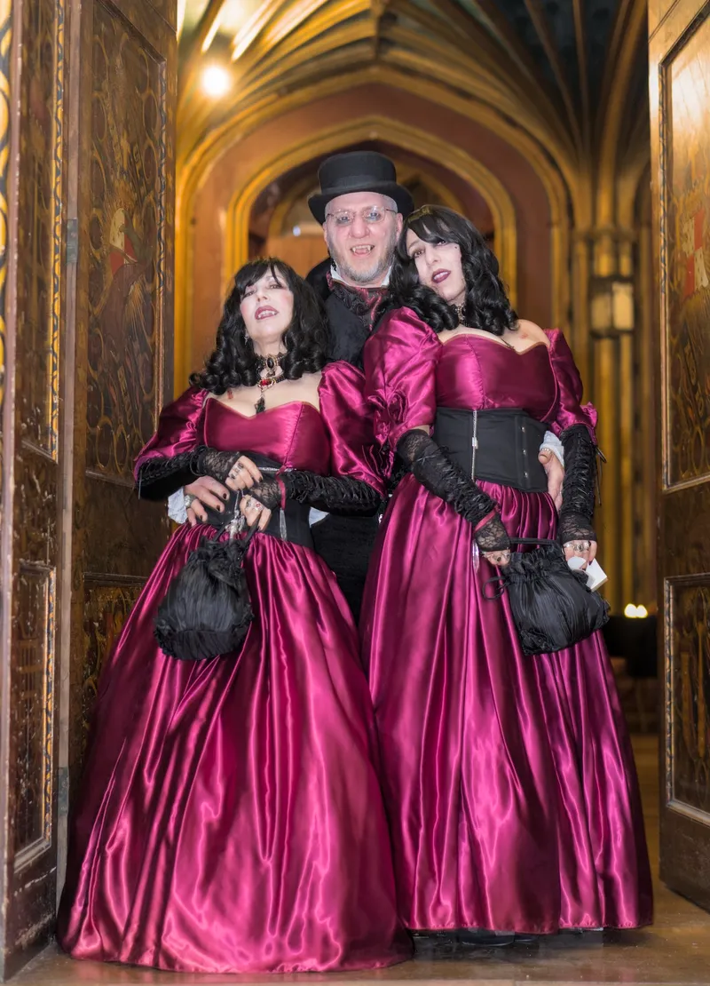 Three individuals pose together in a gothic setting, with ornate architecture visible in the background. The two women on either side wear elegant, deep red dresses with puffed sleeves and black accessories, while the man in the center sports a black outfit with a top hat and glasses. Their attire reflects a vampire theme, fitting the atmosphere of the Vampire Games Day II event.
