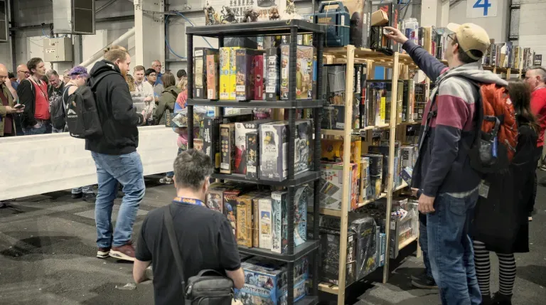 A bustling scene at a tabletop gaming event, featuring a tall shelving unit filled with various board games and miniatures. Attendees are engaged in browsing the games, with one person reaching for a game on the top shelf while others stand in line nearby. The venue is spacious, with a crowd of people in the background, indicating a lively atmosphere.