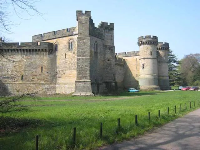 A large stone castle with multiple towers and thick walls, set against a clear blue sky. The castle features a mix of architectural styles, with rounded towers and crenellated parapets. In the foreground, a grassy area is bordered by a low fence, and a few cars are parked nearby.