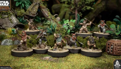 A group of four Ewok miniatures from the Star Wars: Legion game is displayed on circular bases, set against a lush, green forest backdrop. Each Ewok is uniquely posed: one holds a spear, another brandishes a slingshot, while the others are equipped with various weapons and accessories. The terrain features rocks, foliage, and remnants of a crashed vehicle, enhancing the scene's immersive quality.
