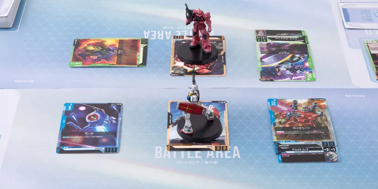 A tabletop scene features two Gundam miniatures positioned on black bases, one in red and white and the other in pink. Surrounding them are several colorful cards displaying various Gundam characters and abilities, with some cards featuring vibrant artwork of mobile suits in action. The play area has a light blue geometric background, enhancing the futuristic theme of the Gundam Card Game.