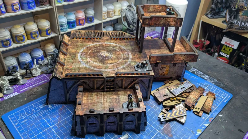 Aged, rustic tabletop terrain piece resembling a fortified stronghold or industrial structure. The main platform has a circular design with a star-like emblem at its center, surrounded by a metal grate pattern. Three arched openings are visible at the base, with two small painted miniatures standing near them—one in green armor and one in black. A second, smaller platform with a ladder leans against the main structure, holding two additional miniatures: one in black and one in metallic silver.

In the background, various hobby supplies are visible on shelves, including bottles labeled "TALI'S MINIATURES COLOURS," "CITADEL," and other paints and tools. A coiled airbrush hose hangs nearby. To the right, a stack of unpainted laser-cut wooden pieces lies on the blue-tiled surface. A small yellow-and-black warning sticker is visible on the side of the main structure.