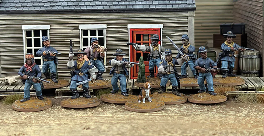 A group of ten detailed 28mm scale miniatures representing dismounted US Cavalry and Buffalo Soldiers stands in front of a wooden building with a red door. The miniatures are posed with various weapons, including rifles and a bugle, showcasing a mix of uniforms and accessories, such as scarves and hats. A small beagle and a cactus are also present among the figures, all mounted on 25mm round bases with a sandy terrain effect.