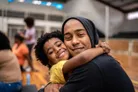 A joyful scene captures a smiling child with curly hair embracing an adult, who is also smiling and wearing a hoodie. They are in a spacious indoor setting, likely a gym or community center, with other people in the background engaged in various activities. The warm expressions and close interaction convey a sense of love and connection between them.