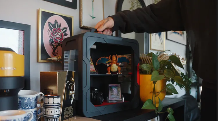 A modular, 3D-printable display case, designed for collectors, is shown in the image. The case features a sleek black design with a snap-fit assembly, and is currently being handled by a person, showcasing its ease of use. Inside the case, various collectibles are displayed, including a Charizard figure, a camera, and trading cards, while decorative items and framed artwork are visible in the background.