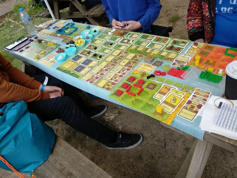A tabletop game setup is displayed on a light blue table, featuring a variety of colorful game pieces and cards. The game board is filled with hexagonal tiles, tokens, and cards in shades of green, yellow, red, and brown, indicating different resources and actions. In the foreground, a person wearing an orange sweater and black pants is seated, with a blue backpack visible beside them, while another player’s hands are seen above the game board.