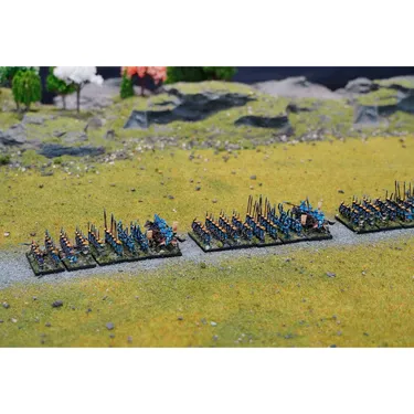 A detailed tabletop scene features numerous 10mm miniatures arranged in formation on a grassy battlefield. The figures include Ashigaru with spears and banners, painted in shades of blue and orange, alongside a few Samurai figures, all positioned on bases along a dirt path. The backdrop includes small trees and rocky terrain, enhancing the setting for Sengoku-era Japan battles.