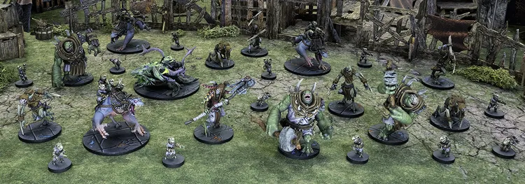 A diverse array of miniatures representing the Urmeck faction from the Maladum tabletop game is displayed on a grassy terrain. The scene includes various creatures, such as smaller Dregs and larger Brutes, alongside Ripclaws ridden by Dregs. The miniatures feature intricate details, showcasing their savage designs and unique poses, set against a backdrop of wooden structures and overgrown foliage.