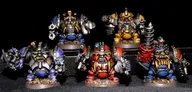 Five multipart, white metal miniatures of armoured dwarves are displayed against a dark background. The front row features two dwarves in vibrant blue and red power armour, each holding different weapons, while the back row showcases three more dwarves in a mix of gold and metallic colours, also armed. Each miniature is intricately detailed, with various mechanical elements and unique designs, highlighting the sci-fi and Victoriana influences.
