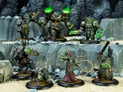 A group of intricately detailed miniatures from the Warmachine tabletop game is displayed on a rocky terrain backdrop. The figures include a large, armored character with green flames emanating from its shoulders, flanked by a menacing wolf-like creature and several other warriors adorned with ornate armor and shields. In the foreground, a female character in a flowing red robe wields a whip-like weapon, while another figure holds a shield and a weapon, showcasing the rich variety of designs and colors typical of the game's factions.