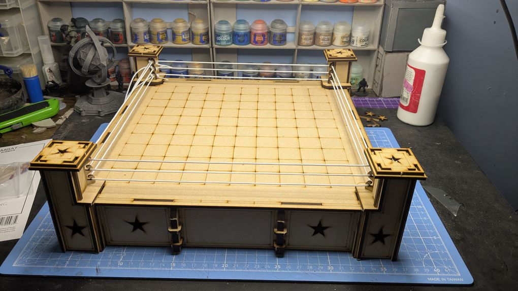 A wooden Rumble Slam Rookie Ring is displayed, featuring a square mat with a grid pattern and four corner posts adorned with star designs. The ring is surrounded by white ropes, and the base has a simple, clean design with additional star motifs. In the background, various paint pots are visible, indicating a workspace for miniature painting and modeling.