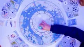 A hand points at a circular Arctic-themed board game map with illustrated wildlife icons, constellations, and geographic markers. Blue-toned background with symbols for animals, regions, and celestial elements.