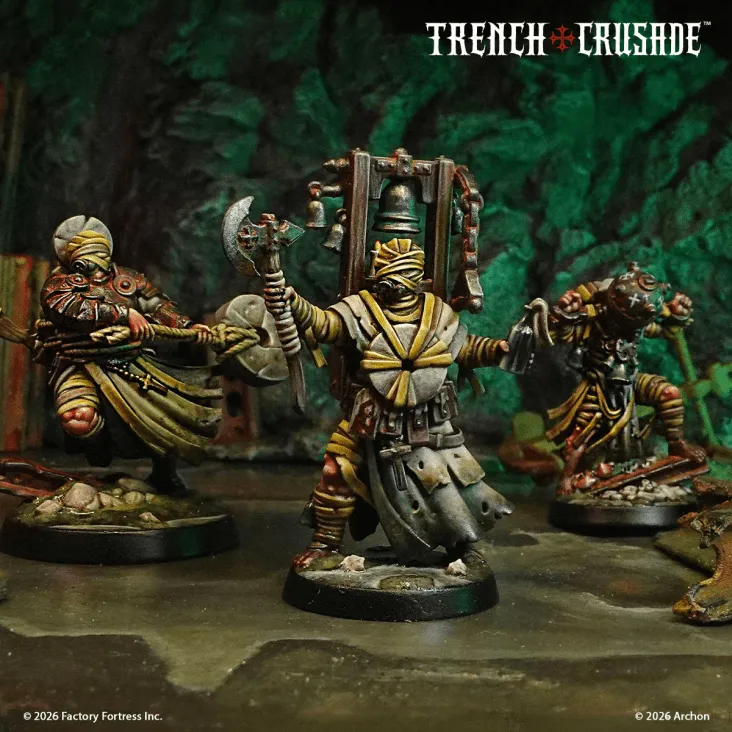 Three intricately designed miniatures from the Trench Crusade game are displayed in a dark, atmospheric setting. The central figure, dressed in tattered robes and wrapped in bandages, wields an axe and carries a large bell on its back, while the other two figures, also wrapped in bandages, are posed with various weapons and equipment. The background features a textured, rocky surface, enhancing the grimdark aesthetic of the scene.