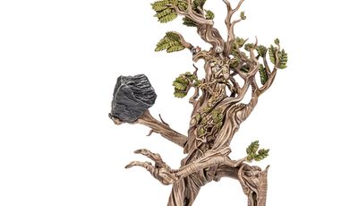 A towering model of Beechbone, an ent from the Middle-earth Strategy Battle game, stands prominently with a rugged, bark-like texture and intricate details. His limbs are gnarled and branch-like, adorned with vibrant green leaves, while he holds a large rock in one hand. The base features rocky terrain with patches of grass, emphasizing his formidable presence on the battlefield.
