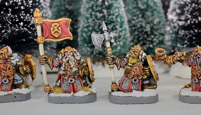 Three painted miniature figures of fantasy dwarves stand on round bases in a snowy outdoor setting with blurred evergreen trees in the background.

The left figure wears a horned helmet, patterned tunic, and fur cloak, holding a pole-mounted red banner with a gold emblem of a snarling face. The figure also carries a round shield on their back.

The center figure, slightly behind the others, wears a helmet with a nose guard, a fur-lined cloak, and carries a pole weapon with a curved blade.

The right figure, holding a large double-bladed axe over one shoulder, wears a horned helmet, fur cloak, and patterned tunic. Their beard is braided and wrapped around their waist.