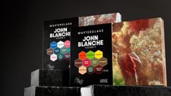 Two sets of John Blanche Masterclass paints are displayed, with Volume 3 on the left featuring a cool color palette, including shades like "Farewell" and "Shadow Thorn," and Volume 4 on the right showcasing warm tones such as "Emberowl" and "Blood Thorn." Both sets are presented in sleek black packaging with vibrant hexagonal labels indicating the paint colors. The background is dark, enhancing the visual impact of the paint sets.