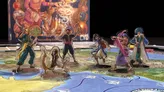 Five character standees are positioned on a colorful game board depicting a map inspired by Arabian Nights. The characters include a seated figure in white robes, a man in a red vest and black pants, a woman in a flowing pink outfit, a man in a blue vest with an orange satchel, and an older man in a blue turban and white robe. In the background, the game box is partially visible, featuring vibrant artwork and the text "PLAY." The board includes various paths and locations, hinting at the adventures players will embark on.