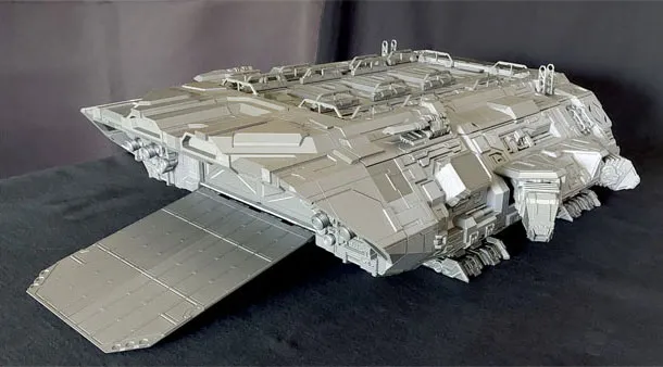 A large, detailed model of a spaceship, featuring a flat, ramp-like structure at the front and a complex array of panels and textures across its surface. The ship has a robust, angular design with various protrusions and openings, suggesting functionality for cargo or troop deployment. The model is presented in a light gray color against a dark background, highlighting its intricate details.