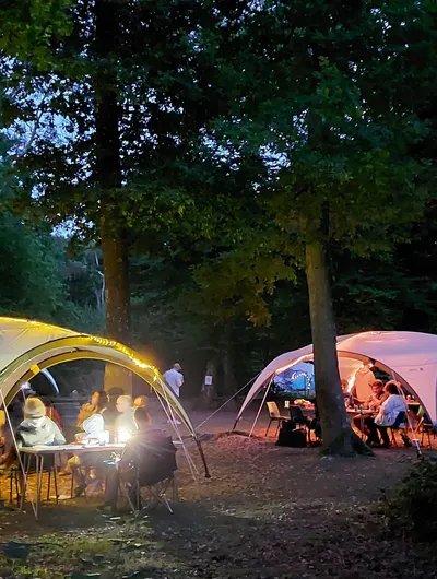 Two large, white canopies are set up in a wooded area during dusk, illuminated by warm lights. Under one canopy, a group of people sits around tables, engaged in conversation and activities, while another canopy hosts a similar gathering. The surrounding trees create a serene atmosphere, enhancing the friendly and inclusive vibe of the event.