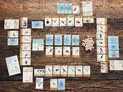 A wooden surface with numerous cards arranged in rows, each depicting illustrations of birds, eggs, food tokens, and various game symbols. Some cards have blue or light backgrounds with circular food tokens in green, brown, and yellow. A cluster of small pink and white egg miniatures is visible near the center-right. The cards display bird names, habitat types, and numerical values. The layout suggests a board or card game in progress.