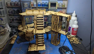 A detailed assembly of modular terrain pieces made from laser-cut wood, featuring multiple levels, staircases, and platforms. The structure includes various textures and patterns, with a mix of open and enclosed areas, showcasing a futuristic industrial design. In the background, paint bottles and tools are visible, indicating a workspace for painting and customizing the terrain.