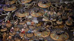 A detailed tabletop scene features various miniatures from the Middle-earth Strategy Battle Game, depicting a tense moment in Mirkwood. Prominent are several large spiders on circular bases, surrounded by a mix of characters including Bilbo, Thorin, and other dwarves, all positioned on a textured terrain with rocks and twisted roots. The dark, earthy colors of the setting enhance the atmosphere of the iconic "Flies and Spiders" chapter from The Hobbit.