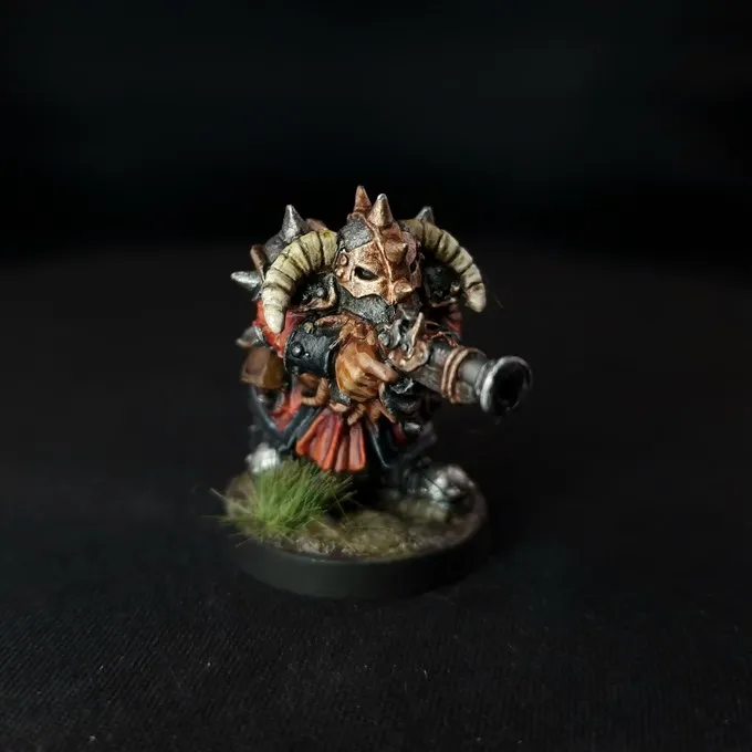 A painted miniature figure of a Chaos Dwarf with a bull-like head, curved horns, and a beard. The figure wears dark armor with orange and red fabric details, and holds a cannon-like weapon on its right shoulder. The base has green grass-like texture. The background is black.