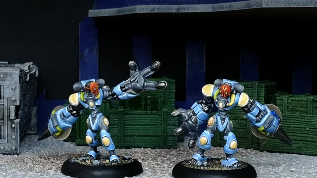 Two Assault Androsynth miniatures are prominently displayed in the foreground, designed for hand-to-hand combat. They feature a blue and yellow color scheme, with one holding a Vibro Scythe and the other a Cesti, showcasing their imposing, armored forms. The background includes dark crates and a textured surface, enhancing the urban warfare theme.