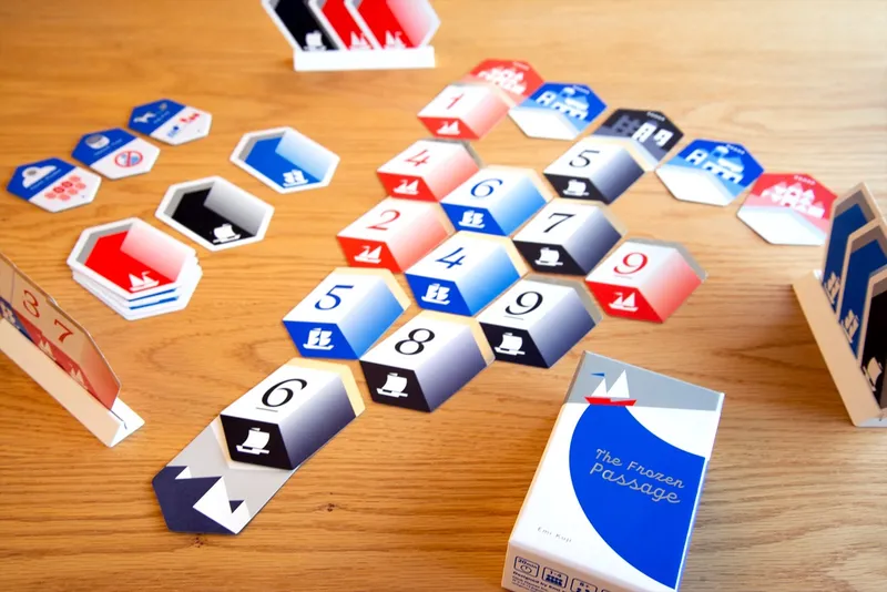 A tabletop scene features various hexagonal game tiles in red, blue, and black, arranged on a wooden surface. The tiles display numbers and symbols, including boats and icons, indicating gameplay elements. In the foreground, a card holder displays a card with the number "37," while a box labeled "The Frozen Passage" is positioned to the right, showcasing its colorful design.