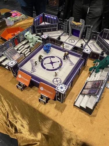 A detailed tabletop gaming arena is showcased, featuring a wrestling ring with a purple star emblem at its center. Various miniature figures, including a blue creature and a green one, are positioned around the ring, while stands and steps are visible on the sides. The arena is adorned with vibrant decorations and signage, with branding from 