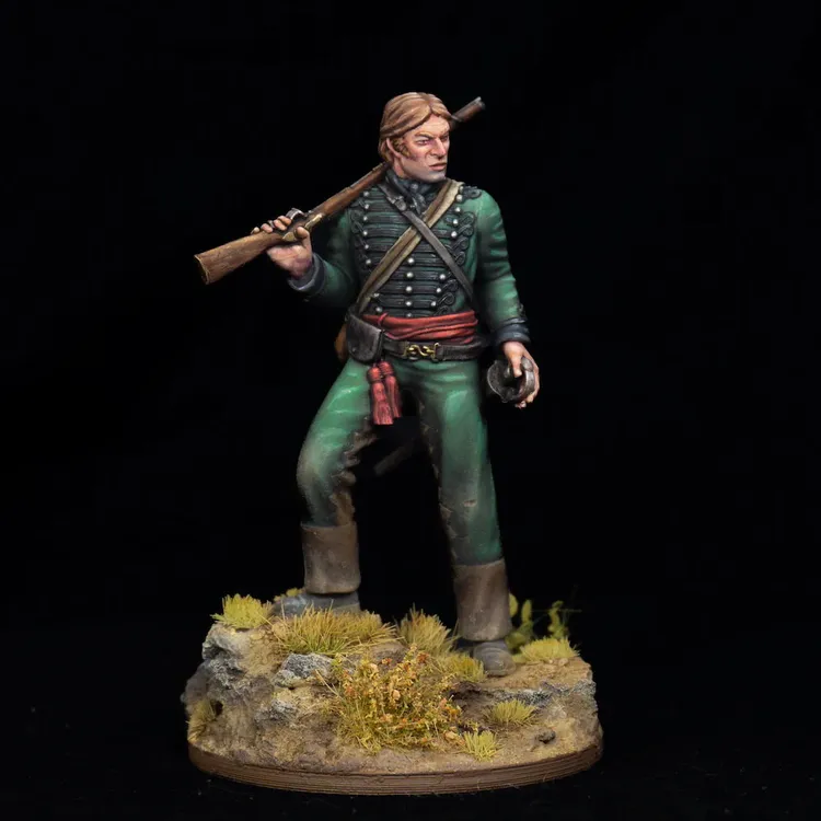 A detailed miniature of Richard Sharpe stands confidently, holding a rifle over his shoulder and a pistol in his right hand. He is dressed in a green military uniform with black buttons and a red sash, set on a textured scenic base featuring rocky terrain and tufts of grass. The figure captures a dynamic pose, showcasing intricate details in the clothing and facial expression.