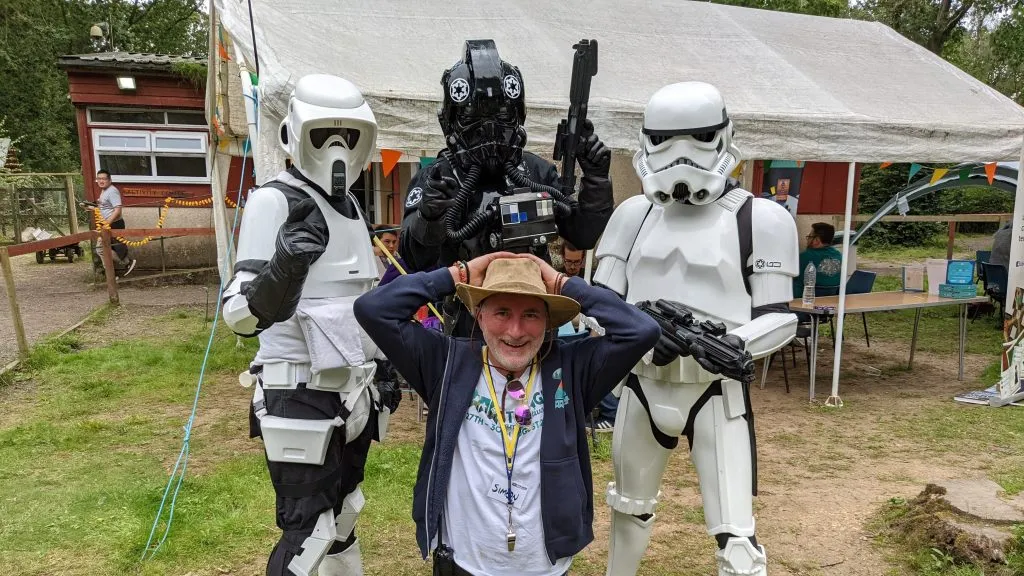 A man in a wide-brimmed hat and a casual outfit kneels in the foreground, smiling and playfully raising his hands above his head. Behind him, two Stormtroopers and a TIE Fighter pilot in full costume stand with their weapons, creating a fun and engaging atmosphere. The setting features a grassy area with a tent in the background, suggesting a lively outdoor event.