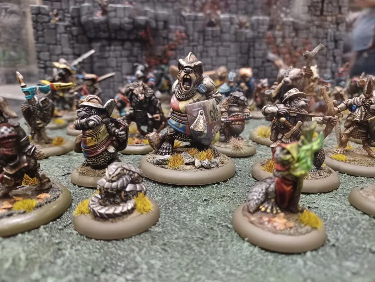 A group of anthropomorphic animal miniatures, primarily featuring badger-like figures, is depicted in a dynamic scene. The miniatures are intricately painted, showcasing a variety of colors and details, with some wearing armor and wielding weapons, while others appear to be casting spells or holding shields. The background features a textured terrain with grass and rocks, enhancing the fantasy setting, and a stone wall is visible in the distance, adding depth to the scene.