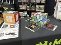 A wooden cube puzzle game on a black table with six colored cube types—forest, ocean, cave, desert, lava, tundra—partially assembled. Two game boards with numbered tiles and a red scoring token lie nearby. Shelves in background display other board game boxes.