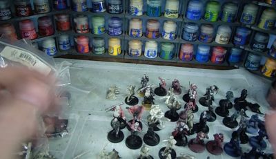 A variety of Heresy Miniatures models are arranged on a work surface, showcasing a mix of painted and unpainted figures in various poses. In the background, shelves filled with paint pots in a range of colors, including blues, greens, and reds, provide a vibrant backdrop. The models include soldiers and fantastical characters, some with detailed bases, while the surface beneath them is marked with paint splatters and tools of the hobby.